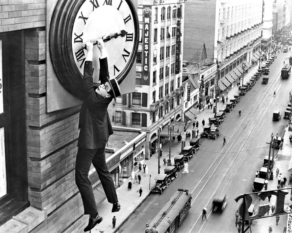 A man is hanging from the hand of a clock fixed on the exterior wall of a six-story building, risking his life.